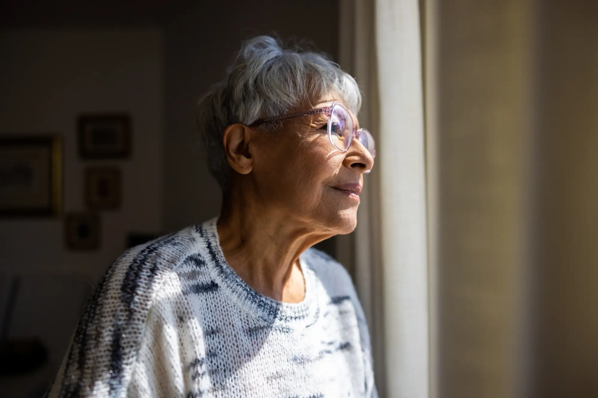 A beautiful multiracial senior woman looking outside the window in her home at The Delaney at The Vale in Woburn, MA.