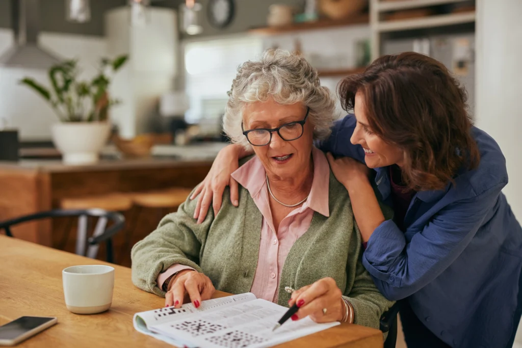 middle age daughter leans over her elderly mother, helping her do a crossword puzzle at the kitchen table