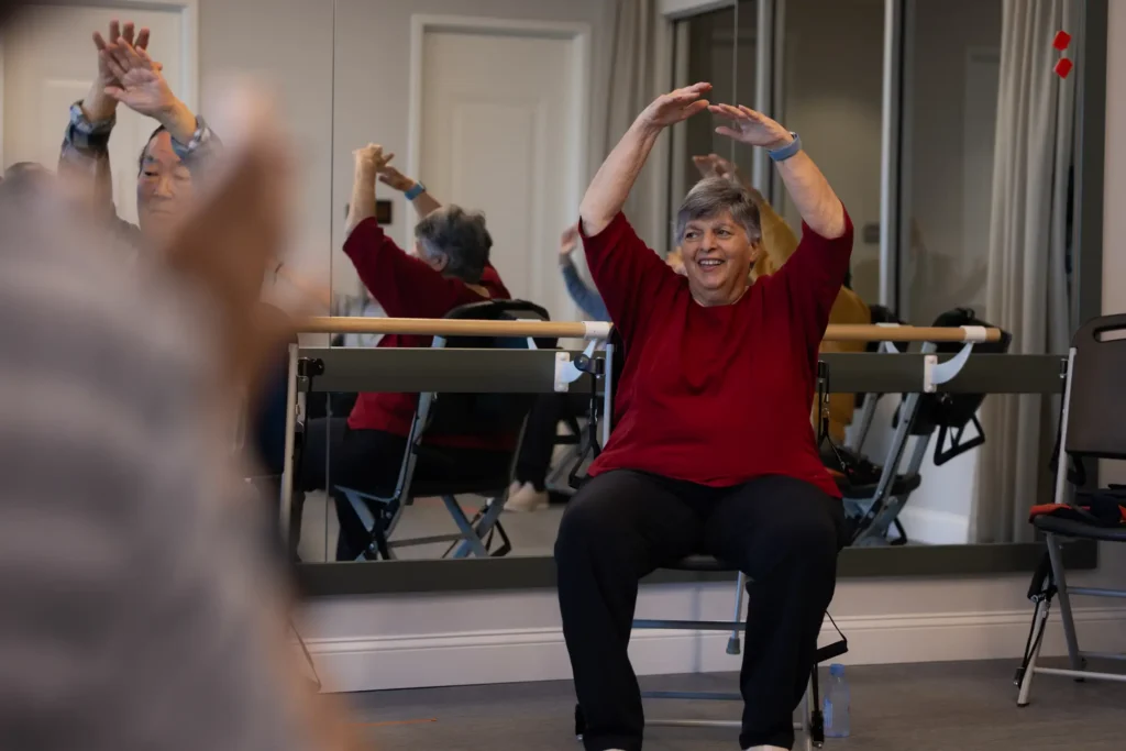 older woman with short gray hair, wearing maroon top and black pants, sits in a chair, holding arms over her head, participating in chair yoga, one of the best activities for seniors with limited mobility at The Delaney at the Green