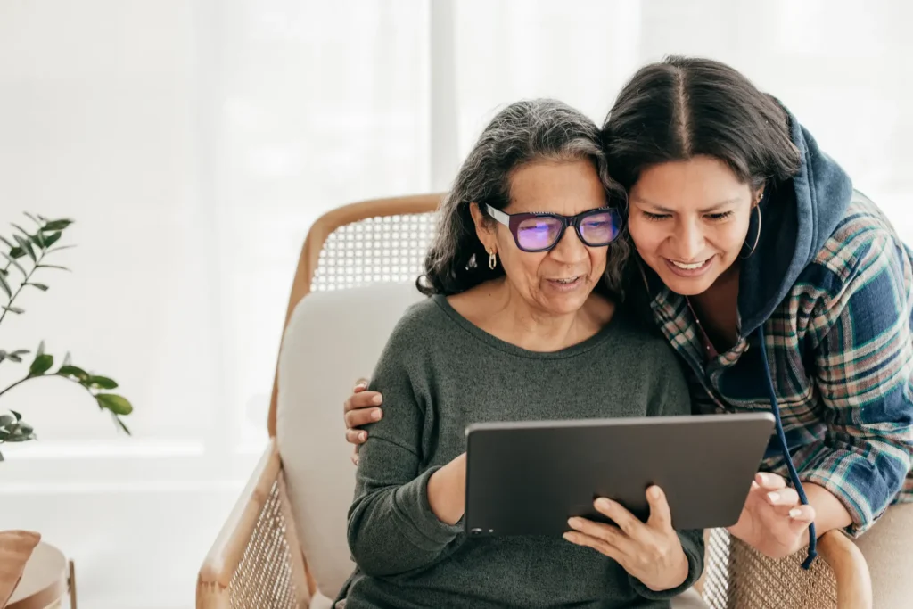 older woman with dark shoulder length hair, wearing glasses and grey shirt, sits in a chair and looks at a laptop held by a younger woman with dark long hair, leaning next to her