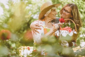 senior woman with white hair wearing a straw hat and holding a basket of flowers is embraced by a younger woman with long brown hair, helping her garden.