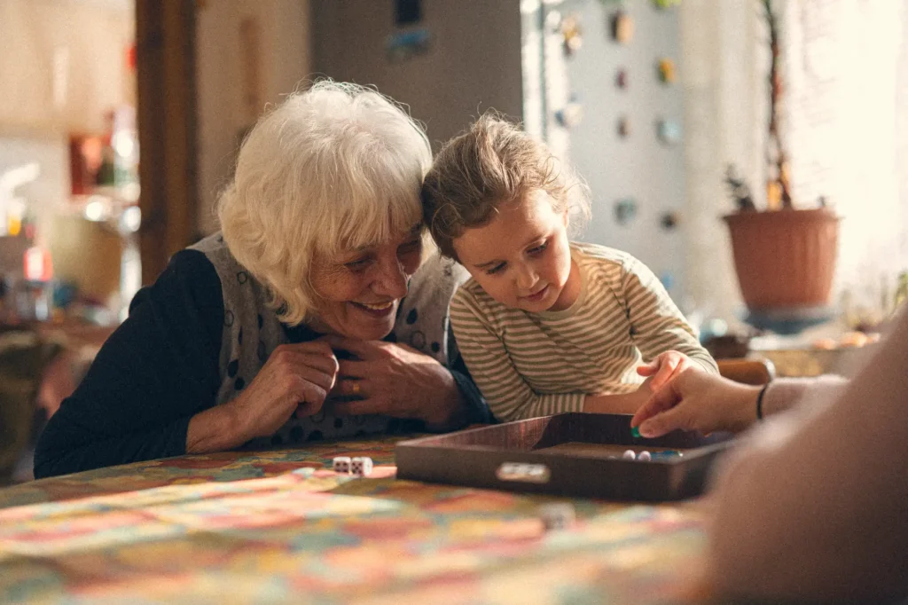 senior woman with white hair sits at table with her young grandaughter, their heads touching as they play a board game, one of the best activities for seniors with limited mobility.